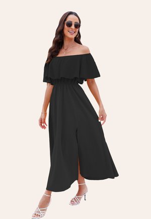 Zwarte maxi jurk zonder schouders met ruches op de mouwen, elastische taille en hoge split tot de bovenbenen; gedragen met witte gespenhakken en een zonnebril.