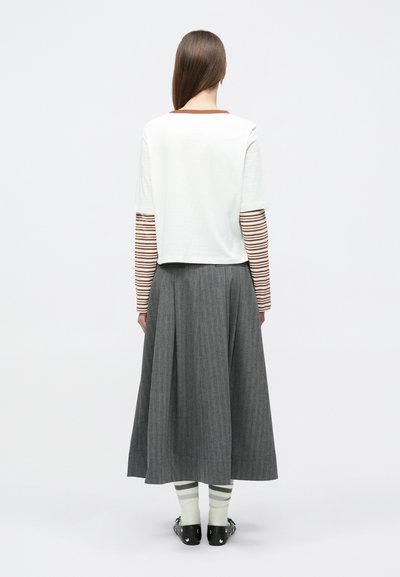 Λευκό κοντό τοπ με ριγέ μακριά μανίκια, γκρι πλισέ φούστα και ανοιχτοπράσινες κάλτσες με σκούρες ρίγες, φορεμένα με μαύρα παπούτσια χωρίς κορδόνια.