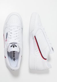 White leather sneakers with red and navy accents, featuring perforations on the toe, laces, and a logo on the side and tongue.