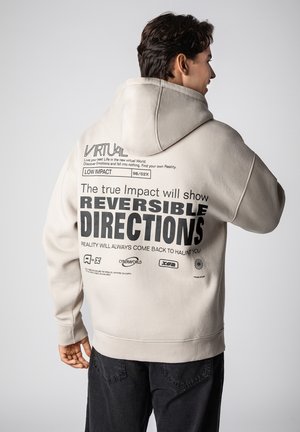 Beige hoodie with a drawstring hood. Features large black text on the back, including "REVERSIBLE DIRECTIONS," with various small logos below.