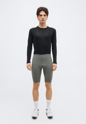 Giovane uomo che indossa una maglia da ciclismo nera a maniche lunghe, pantaloncini imbottiti grigi con bretelle, calzini bianchi e scarpe da ciclismo bianche, in piedi dritto.