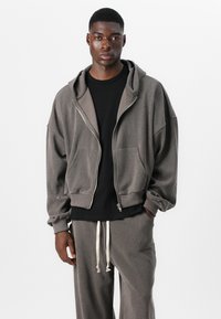 Gray zip-up hoodie with a textured fabric, large front pockets, and a relaxed fit. Paired with dark t-shirt and gray trousers with drawstrings.