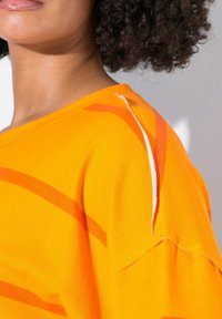 T-shirt arancione brillante a maniche corte, con strisce diagonali arancioni più chiare e bianche, e cuciture visibili sulla cucitura della spalla.