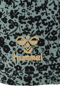 Blommönstrad tyg i grönt med svarta blommor. Har en guldig logotyp och detaljer, med en mjuk, texturerad yta.