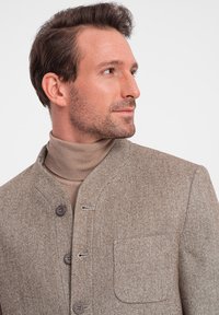Braune Fischgrätjacke mit Stehkragen, Knopfverschluss und einer Brusttasche. Getragen über einem hellbeigen Rollkragenpullover.