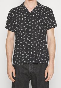 Maglietta nera a maniche corte con stampe grafiche bianche di vari simboli; presenta un colletto a revers e una chiusura con bottoni.