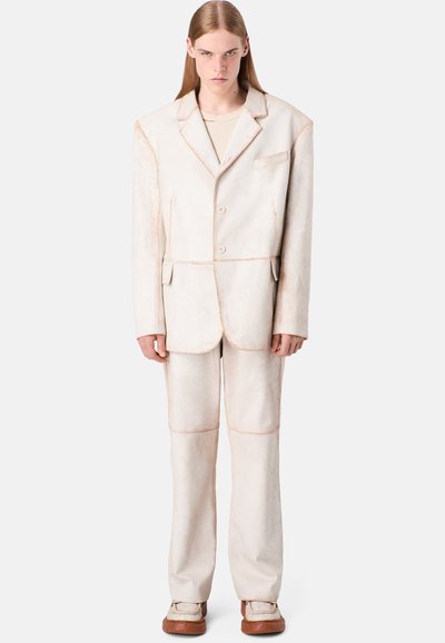 Beige tailored suit with visible stitching details, featuring a single-breasted jacket, straight pants, and a slight sheen.