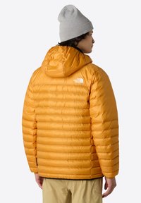 Páperová bunda v živé oranžové barvě, má kapuci, prošívaný design a logo na zádech. Vyrobena z lehkého a hladkého materiálu.