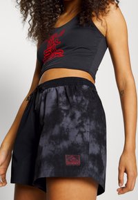 Černý krátký top s červeným grafikou, doplněný černými kraťasy s batikovaným vzorem, elastickým pasem a nášivkou s logem na lemu.