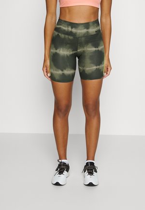 Frau trägt grüne Batik-Athletikshorts, rosa Crop-Top, weiße Sneakers, steht vor einem schlichten weißen Hintergrund.
