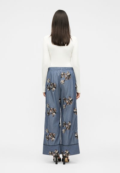 Femme aux cheveux bruns et raides portant un haut blanc à manches longues et un pantalon bleu ample à motifs floraux, debout de dos sur fond blanc.