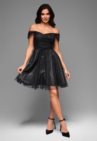 Robe noire décolletée avec corsage froncé, jupe en tulle et subtil éclat. Associée à des chaussures à talons noirs.