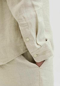 Camisa de lino beige claro con puños puntiagudos y dos botones, bolsillo lateral y textura visible del tejido entrelazado.