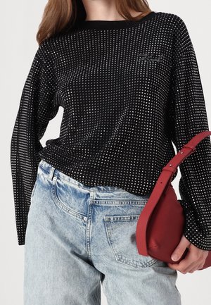 Femme portant un haut noir à manches longues avec de petits pois argentés, un jean bleu clair et tenant un sac à main rouge avec une sangle autour du poignet.