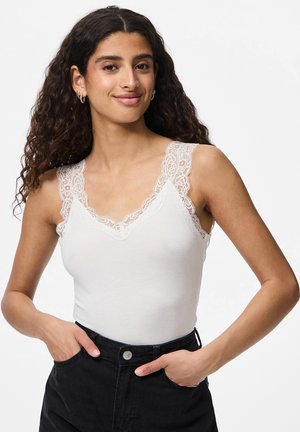 Femme aux cheveux bouclés et foncés, vêtue d'un débardeur blanc bordé de dentelle et d'un pantalon noir, debout les mains dans les poches devant un fond blanc.