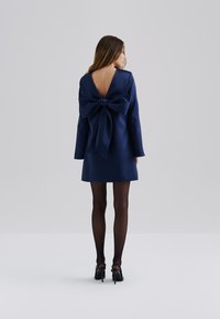 Robe satin bleu marine avec un dos en V profond et un grand nœud à la taille. Des manches longues et une silhouette droite complètent le design.