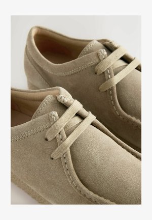 Béžové semišové boty s kulatou špičkou, texturovaným povrchem a plochými tkaničkami. Obsahují tenké švy podél okrajů a nízkoprofilovou podrážku.