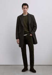 Cappotto di lana scuro con ampie risvolte, indossato sopra un maglione verde oliva, abbinato a pantaloni verde scuro e stivali in suede marroni. Design semplice e sartoriale.