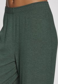Zaļas ribotas auduma bikses ar elastīgu jostu, ar fakturētu virsmu un vaļīgu piegriezumu, daļēji atklājot krokas sānos.