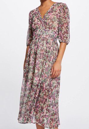 Vrouw draagt een doorschijnende midi-jurk met bloemenprint, pofdriekwart mouwen en een V-hals tegen een effen witte achtergrond.