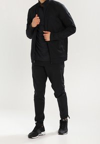 Černá sportovní bunda s vysokým límcem, v kombinaci s černým topem a sportovními kalhotami. Texturovaná tkanina, kapsy na zip a sportovní obuv.