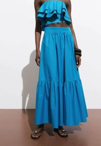 Modri bombažni komplet, ki vključuje ruffle crop top in pliseirano maksi krilo. Krilo ima elastičen pas in gladko teksturo.