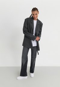 Grå strukturert blazer med løs passform, parret med mørke denimbukser og hvite joggesko. Blazeren har en snittet krage og knyte-detalj.