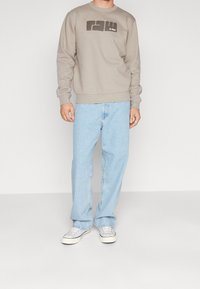 Felpa grigia con logo, vestibilità rilassata, colletto rotondo, abbinata a jeans in denim azzurro chiaro e sneakers in tela grigia. Sfondo semplice.