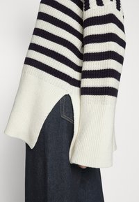 Striped sweater with navy and cream colors, featuring ribbed texture, side slits, and a relaxed fit. Paired with dark denim jeans.