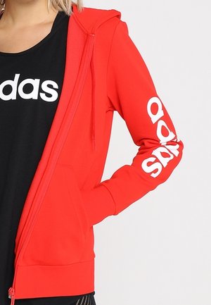 Red zip-up hoodie with white logo on the sleeves, made of textured fabric with a hood and front pockets. Black shirt underneath.
