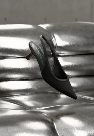 Sort spids tå slingback-sko med lav hæl, der hviler på en skinnende, metallisk sølv tuftet læderoverflade.