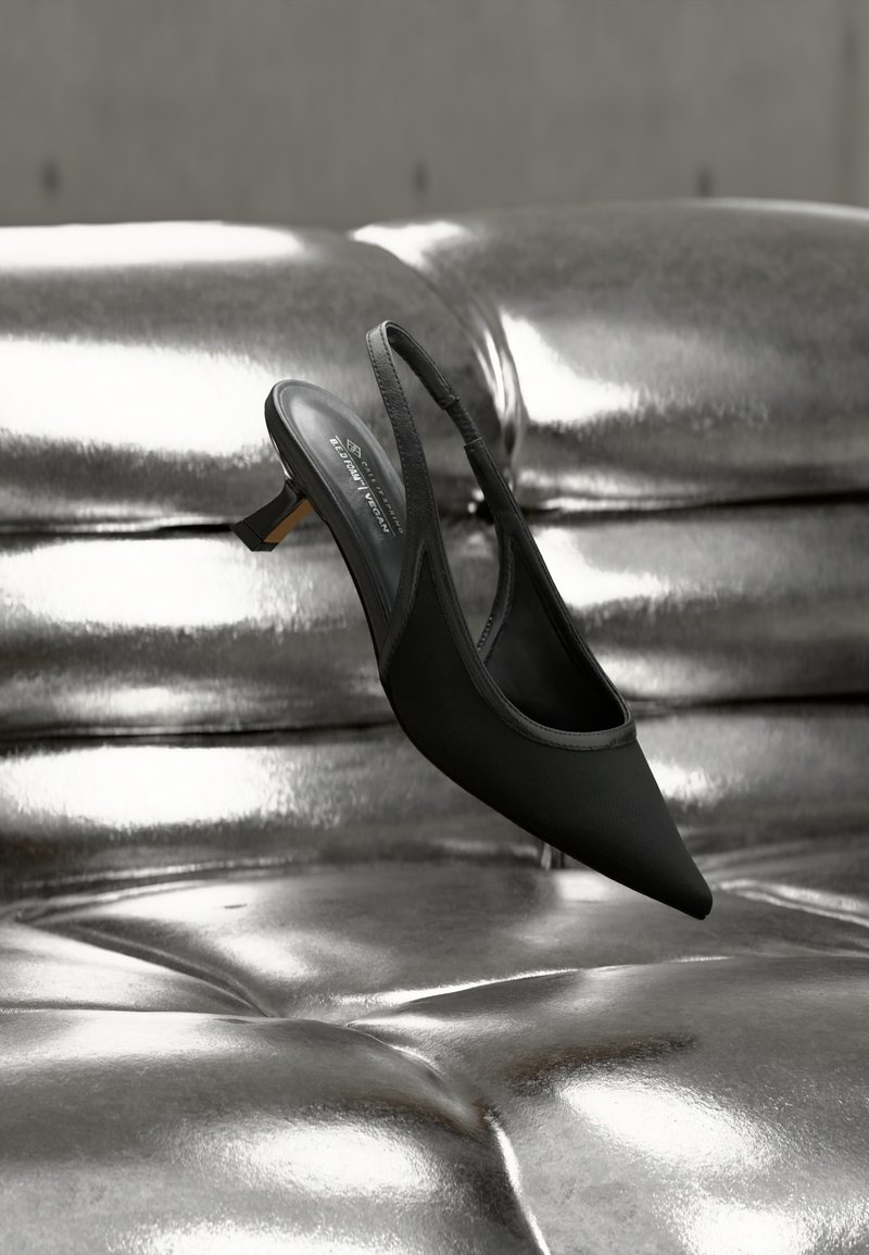 Chaussure noire à bout pointu avec bride arrière et petit talon, posée sur une surface en cuir matelassé argenté métallique et brillant.