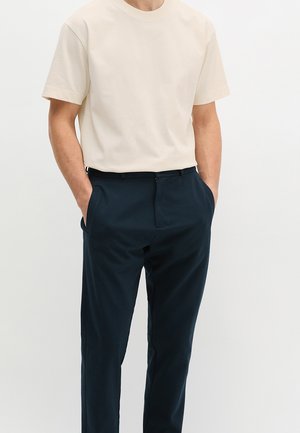 Uomo che indossa una t-shirt crema a maniche corte infilata in pantaloni blu navy su misura, in piedi con le mani in tasca contro uno sfondo bianco.