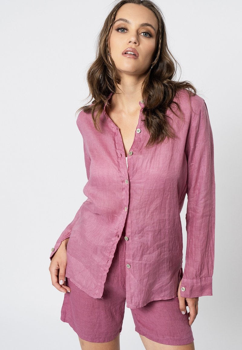 Camicetta rosa in lino con bottoni e maniche lunghe, con orlo leggermente curvo, abbinata a pantaloni corti coordinati, entrambi con una texture informale.