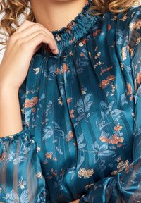 Tealfärgad blus med blommönster, plisserad design och elastisk halsringning och ärmslut. Genomskinligt tyg med subtil lyster och finrandigt mönster.