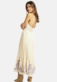 Cremfarbenes, fließendes Maxikleid mit gewelltem Saum, lila floralem Stickereimuster am Saum und verstellbaren Trägern, getragen zu beigen, hohen Stiefeln.