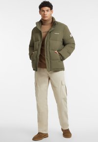 Olivengrøn pufferjakke med høj krave, lynlås foran og sidelommer. Sammen med en brun turtleneck og beige cargo-bukser, der har brune støvler.