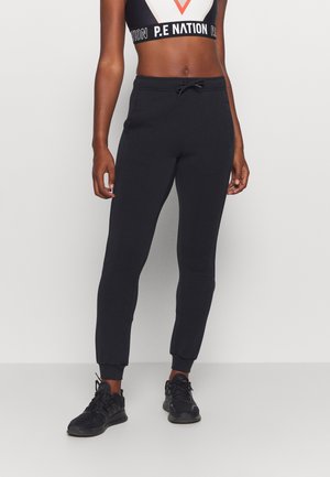 Kvinde iført sorte joggingbukser med snøre i taljen, sorte sneakers og en sort-hvid sports-bh med "P.E NATION" branding.