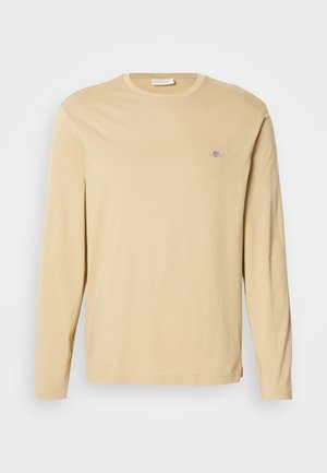 Tricou bej cu mânecă lungă și guler tip crewneck, cu un mic logo brodat pe partea superioară stângă a pieptului, prezentat pe un fundal alb simplu.