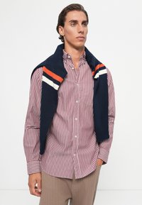 Gestreepte rood-witte shirt met knoopsluiting op de kraag, gedragen onder een marineblauwe trui met oranje en witte strepen op de schouders. Bruine gestructureerde broek.
