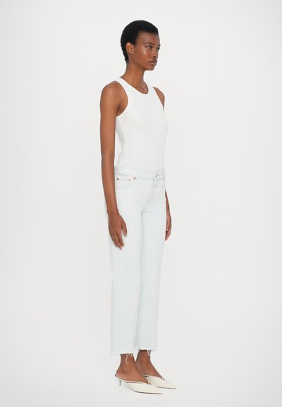 Top de tirantes blanco con canalé combinado con jeans de talle alto azul claro con dobladillos deshilachados, llevados con mules de puntera puntiaguda, de pie contra un fondo blanco.