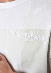 Camiseta blanca de algodón con texto en relieve "PROJECT X PARIS (MEMBERS CLUB)" y un sutil patrón texturizado en toda la tela.