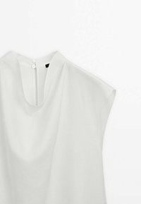 Blouse blanche sans manches avec un col rond et deux petits boutons au col arrière sur un fond uni.