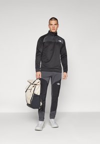 Crna zip-up flis jakna sivo-ružičastih akcenta, uparena s tamnosivim tajicama. Model drži bež i crni ruksak.