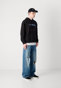 Black hooded sweatshirt with front pocket and logo, paired with light blue distressed wide-leg jeans and black sneakers. Black cap.