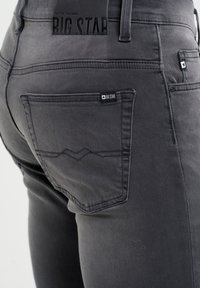 Sivi jeans iz džinsa, ki se prilegajo telesu, z vidno zadnjo žepno torbico z oznako logotipa in diskretnimi detajli šivanja. Vključena je gumijasta oznaka na pasu.