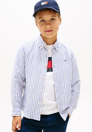 Niño con gorra azul marino, camiseta blanca con logo rojo y negro, camisa de botones a rayas azules y blancas, y pantalones azul marino, posando con una mano en el bolsillo.