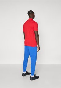 Red polo shirt, blue joggers with elastic cuffs, black athletic shoes, viewed from behind. Features side slits on shirt and white socks.