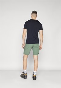 Mørkeblå t-shirt, grønne shorts med sidelommer, og sorte vandresko med orange detaljer. Kort hår, set fra bagenden.