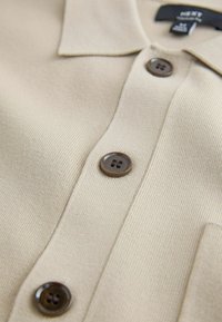 Prim-plan cu o cămașă tricotată bej, cu guler și trei nasturi maro închis vizibili de-a lungul fantei frontale.